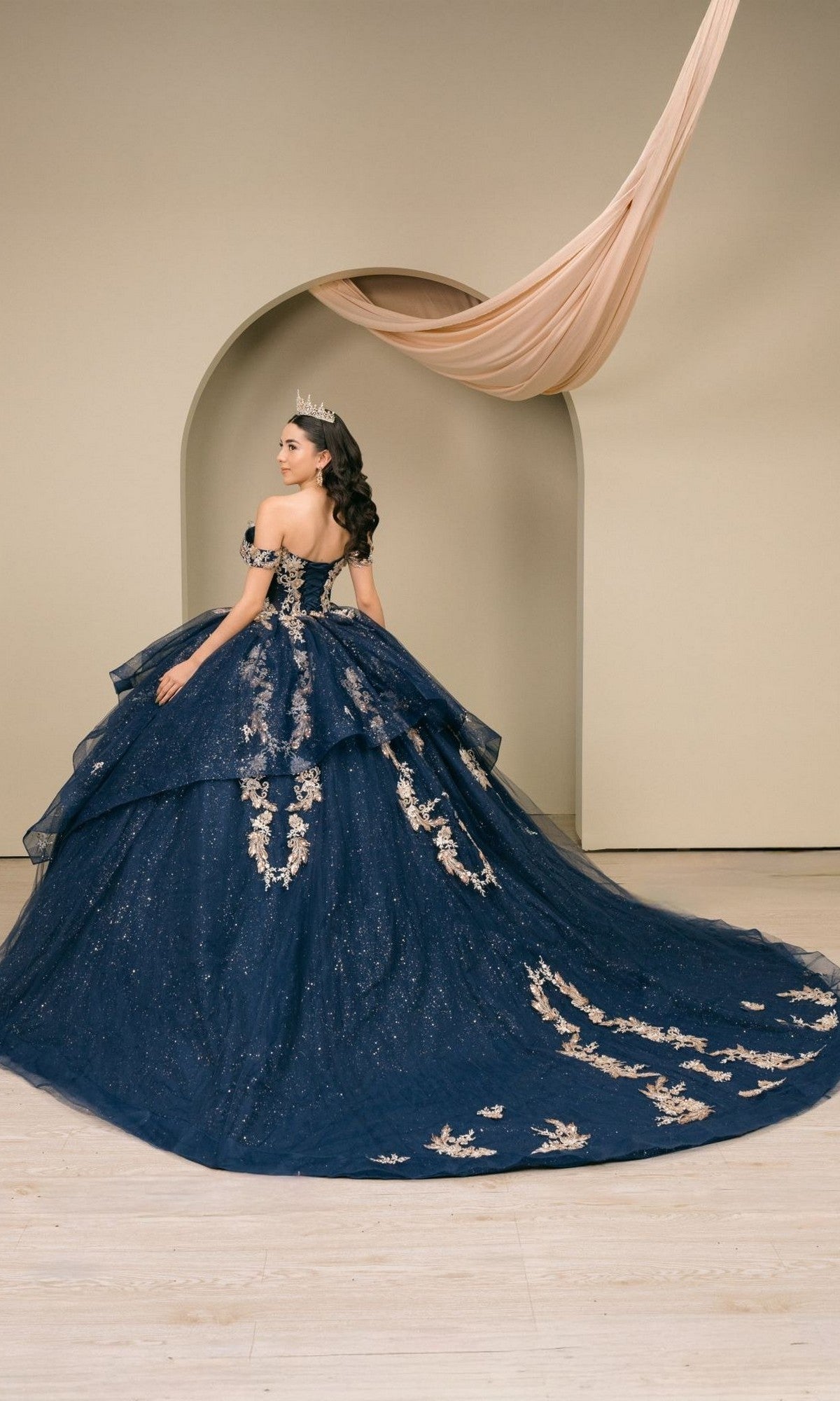 A woman in the Quinceanera Dress 8390JQ by Cinderella Couture—a navy ball gown with gold embroidery and a sweeping train, crowned and poised before a beige archway draped in fabric—radiates timeless Cinderella Couture elegance.